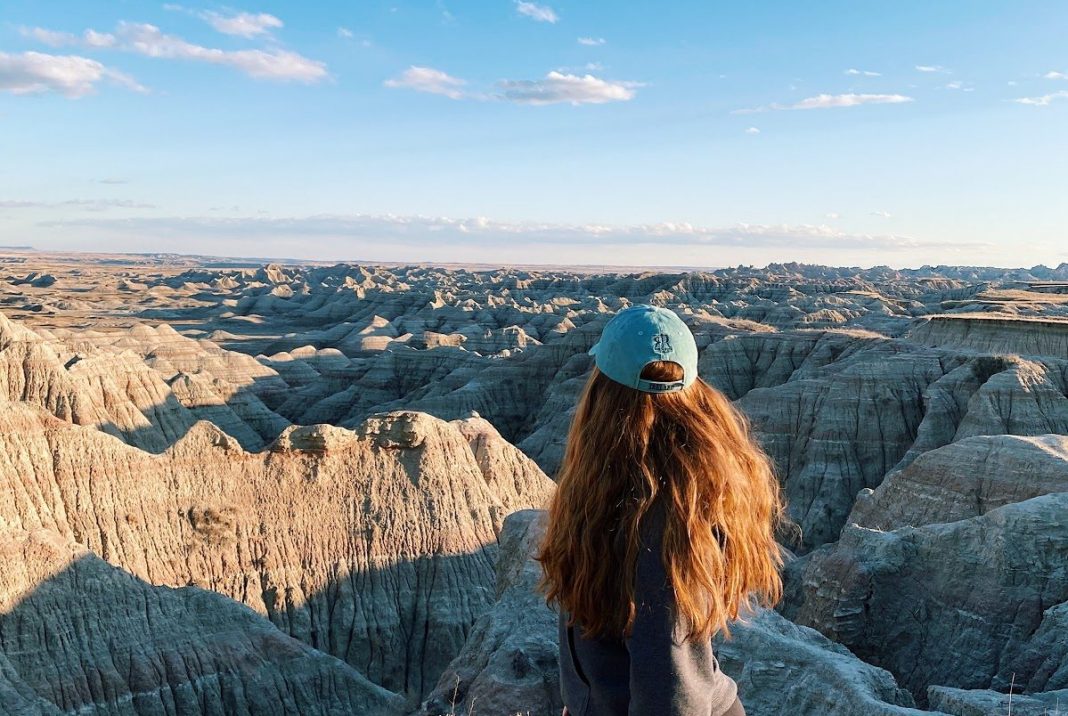 A woman with long red hair stands with her back to the camera. She overlooks a view of rock formations that stretch far into the distance under a blue sky.
